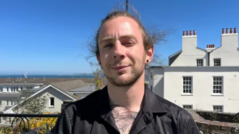Josh Coutu pictured brunette hair tied back. He has a dark, short, beard and moustache. Josh is wearing a black button down shirt and is smiling with closed lips at the camera.