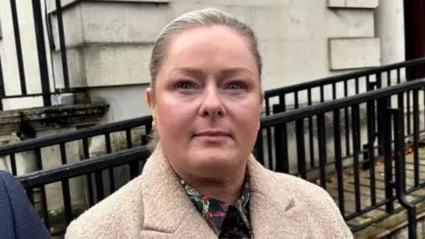 Ann McClure looking at the camera. She is wearing a beige coat and shirt that is green with pink, white and orange pattern. Behind her there is a cream coloured wall with black railings in front of it.