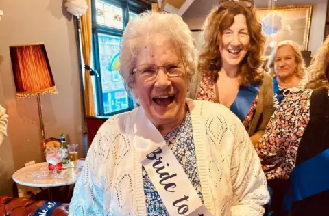 An elderly woman smiles as she wears a "bride to be" sash at her hen do 