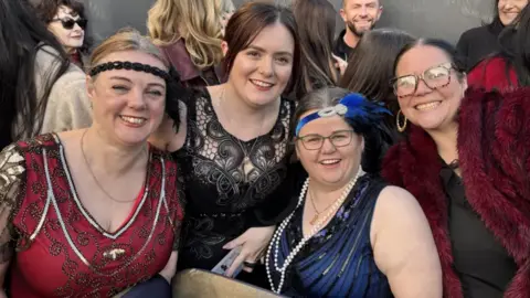 Clair McManus, Susan McManus, Lisa Turner, and Elisha Guiney are each wearing costumes inspired by the early 1900s. They include accessories such as feathered headbands and pearl necklaces. The four women are smiling for the camera and are standing among a large group of people.