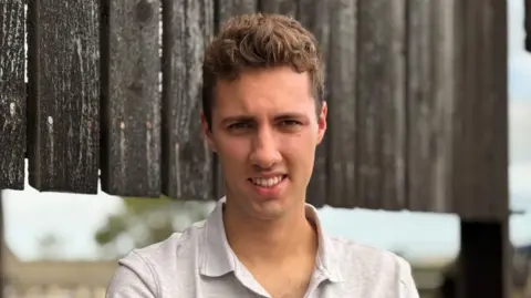 A young man facing the camera. He is wearing a white shirt.