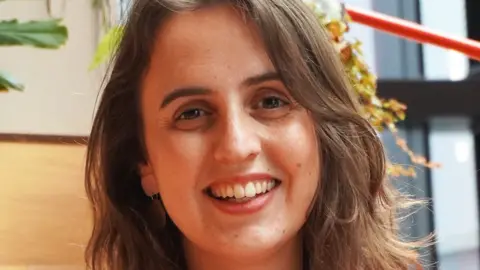 Lucy Jenkins Head shot of Lucy Jenkins, a white woman in her thirties. She has shoulder length layered light brown hair, is wearing light make-up and dangly earrings and is smiling broadly at the camera. She appears to be indoors in a workplace.