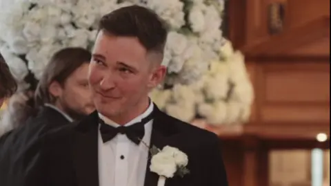 A man with dark brown hair is standing at the end of an aisle. He a black suit jacket, white shirt with black buttons and a black dickie bow tie. He has a white rose pinned to his suit.