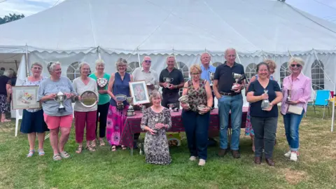 Cheryl Dennis/BBC A line of contestants stand in front of a large marquee. They are holding up various certificates and trophies won at the Bicknoller Flower Show