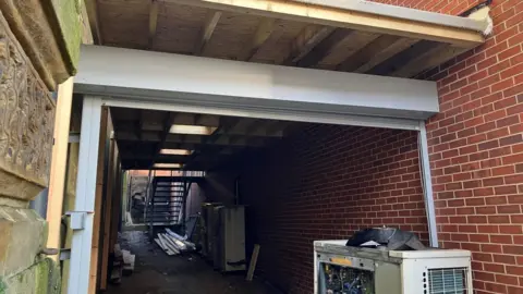 LDRS A newly built roof made of wooden beams and panels is supported by a white steel beam and metal posts. Below it, scattered timber, bricks and other materials cover the ground.
