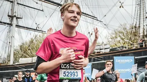 Sportograf / TCS London Marathon A runner in a pink event T‑shirt and race bib is captured mid‑stride during a road race. Tall ship masts and rigging fill the background, indicating a waterfront or historic harbour setting. Spectators line the course holding signs, while sponsor banners are visible along the route.