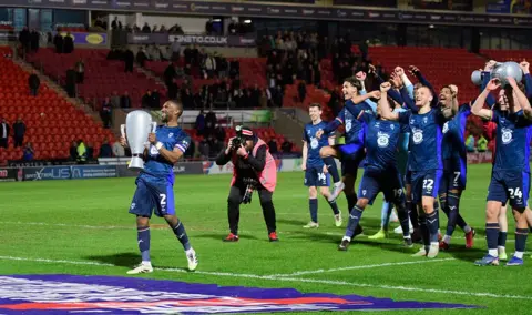 Andrew Vaughan/Getty Images Football players in dark blue kits walk across the pitch carrying a large, silver inflatable trophy. Several players are raising their arms as they walk past a pitchside photographer. Red stadium seating, spectators and advertising boards are in the background under floodlights.