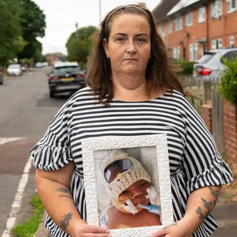 BBC/Chris Waring Hayley Coates, in a striped top, stands on a street holding a picture of Kaylan