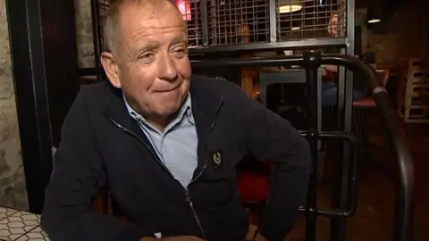 Mick Thurlby with a shirt and zipped top sitting at a table in the bar talking to reporters about the initiative 