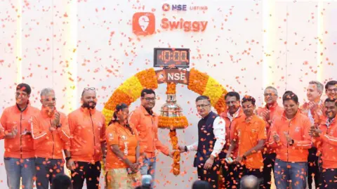Bloomberg via Getty Images Swiggy listing ceremony at the National Stock Exchange in 2024, with confetti falling on Swiggy executives standing on the stage in orange uniforms. 