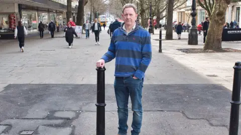 Carmelo Garcia Andrew Booton standing in the middle of the promenade, wearing dark blue jeans and a blue striped jumper. He has short grey curly hair and is looking at the camera with a serious expression. His right hand is resting on top of a black bollard, while his other hand is in his front jeans pocket. Behind him you can see shoppers walking down the road. Beneath his feet there is a large patch of tarmac with has been laid to replace the broken paving stones with cover the rest of the street.