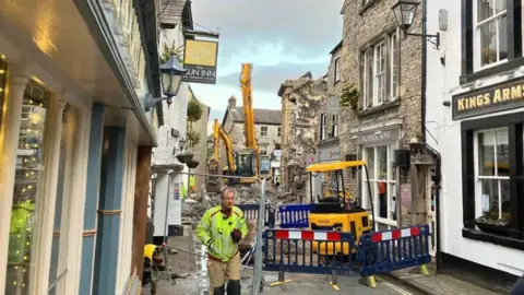 A view down Market Street in Kirkby Lonsdale. The narrow street has been cordoned off with diggers visible. Debris lies in the middle of the street in the distance and a man in hi-vis can be seen in the middle of the photo.