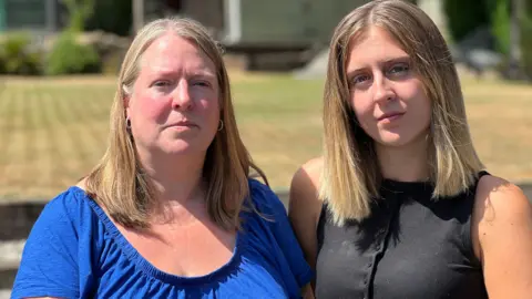 Michelle Lyons/BBC A young blonde-haired woman stand alongside another blonde woman. One is wearing a blue top and the other is wearing a black one.