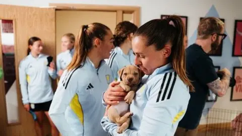 Birmingham Dogs Home A woman in the foreground of the picture has her hair styled in a high ponytail and is dressed in light blue sports gear. She is holding a six-week old puppy and looking down at it. In the background there are four women who are also dressed in sporting clothes and a man in a black tshirt, glasses with tattoos on his arm appears to be holding a camera and looking to the right of the frame. 
