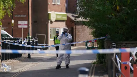 A forensic officer in white overalls standing close to a police cordon taking photographs. There are a number of concrete bollards in view, as well as police tape across the road