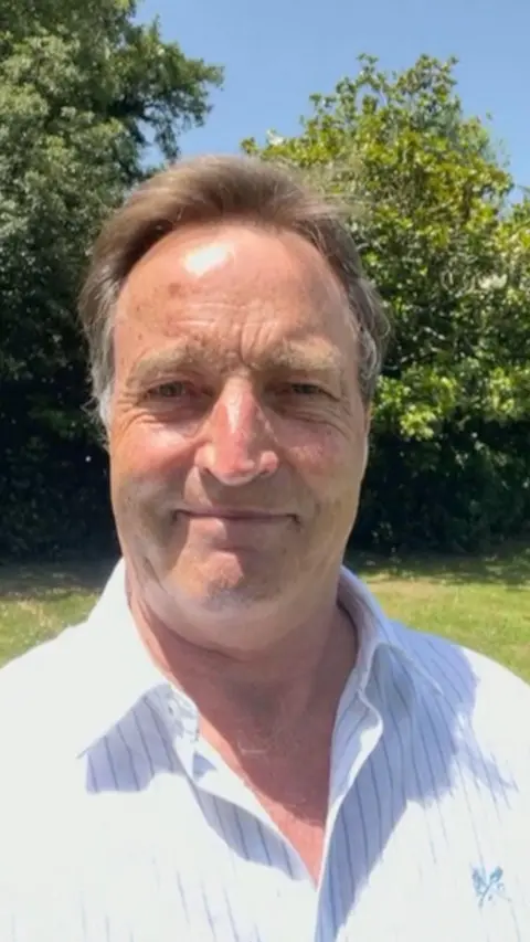 A man with brown hair in a white top. Behind him is green grass and a green tree.