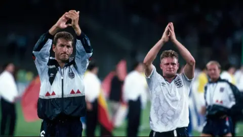 Getty Images Two footballers raise their hands in the air and clap. They wear matching England white football tops and jumpers. Other footballers can be seen behind them. 