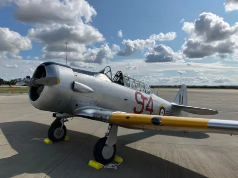 A silver two-seater training aircraft from the 1940s with yellow flashes on the wings, called a T-6 Texan, also known as a Harvard