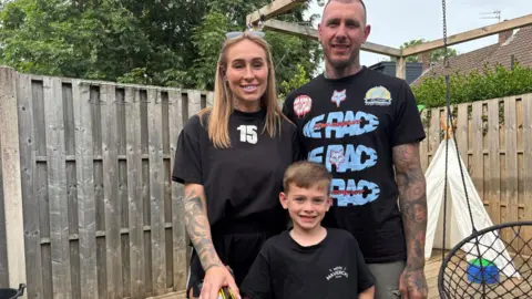 Simon Thake A young boy stands between a man and woman. All three are dressed in black and smiling