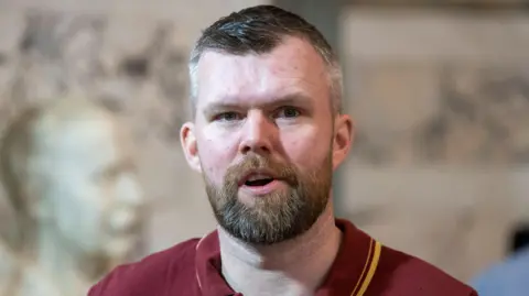 PA WIRE Carroll has short dark and grey hair and a brown beard. He has his mouth open in the picture and is wearing a red polo shirt.