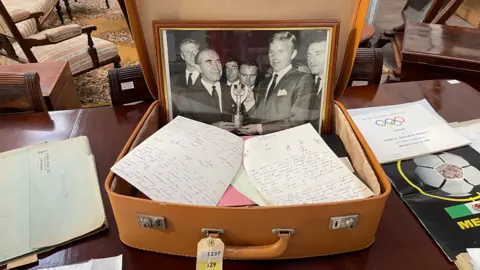 Edd Smith/BBC A brown suitcase with a black and white photograph in a frame, on a brown wooden table with other pieces of papers with writing on it and programmes.