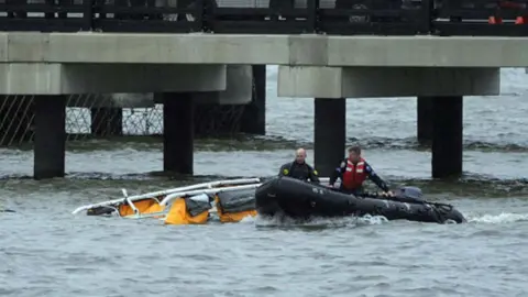 Members of a rescue team are on a black boat near the wreckage of a helicopter crash in a river.