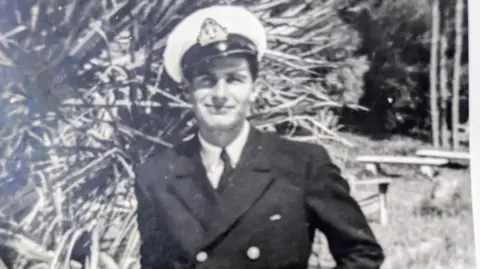 Family Photos A black and white photo of a man in a pilots cap and button jacket. A white collar and tie pokes out the top. He is smiling to the camera.