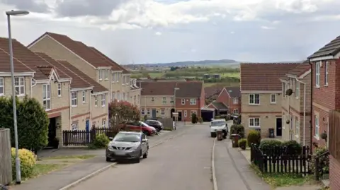 A residential street in Barnsley, with the road a downward slope into the distance. The homes on both sides of the road are relatively new. A hill with trees can be seen in the distance. 