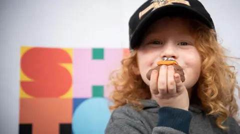 Showtown Blackpool A child with long curly red hair holds a model of fake poo up in line with their face. They are wearing a black hat and grey jumper.