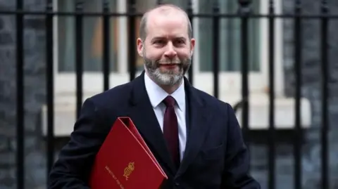 Reuters Jonathan Reynolds, wearing suit and tie and carrying a red folder, is pictured outside in Downing Street, with black railings in the background
