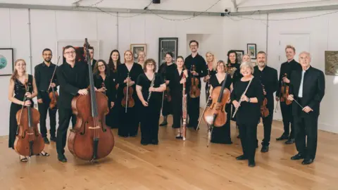 Welsh Chamber Orchestra Seven men and women are dressed in smart black clothing holding string instruments. They are stood in a gallery, with paintings on the white walls and a wooden floor. There are fairy lights hanging from the ceiling.