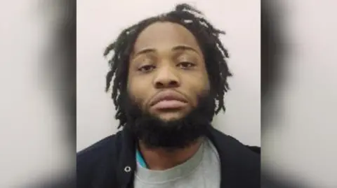 A man in a grey crewneck and black overshirt looks at the camera in a mugshot style image. 