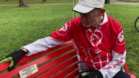 A man in a red and white shirt looks at a red bench with a plaque.