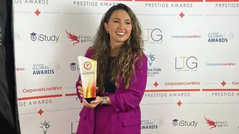 Lissy Cleminson Lissy Cleminson. She is pictured holding an award. She has long dark hair and is wearing a purple suit with a black top. She is smiling, looking to the side of the camera. 