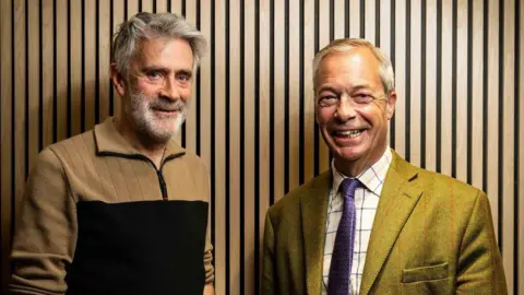 John Nguyen/JNVisuals Christopher Harborne, left, wears a beige and black jumper. He has a full head of grey hair and a grey beard and bushy eyebrows. He stands next to Farage who is grinning. Farage is wearing a green suit and checked shirt and purple tie. 
