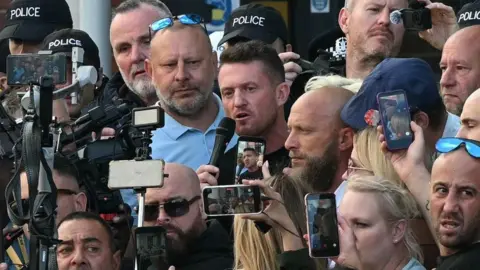 Getty Images Tommy Robinson is pictured speaking to a microphone while surrounded by a large group of people. 