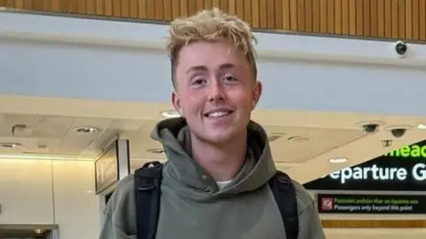 Jamie Online Vintage Jamie Harkin is standing in the airport departures area. He is wearing a green hoodie and wearing a black bag pack. There is an elevator behind him with two people standing outside of it. To his side there are four people with two hugging. In the background there is a large black departures gate.  