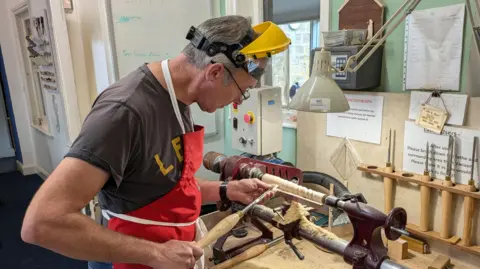 A man wearing a visor and an apron inspects a tool, which bears a resemblance to a large screwdriver, as he stands by a lathe on a workbench.