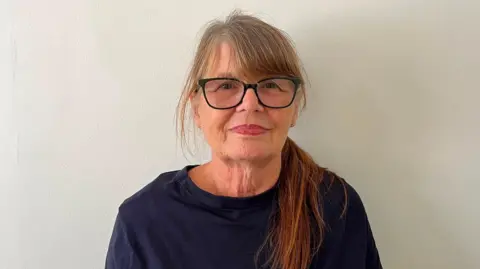 BBC A woman is stood against a white wall looking directly at the camera. She is wearing black glasses and a navy blue shirt. 