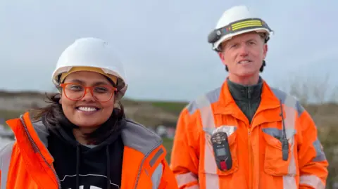 Raj - on the left - smiles at the camera, wearing glasses with an orange frame. She is a young woman with dark hair. Nick - on the right - looks into the camera. He is a middle-aged man. Both wear white hard hats and high-vis orange coats.