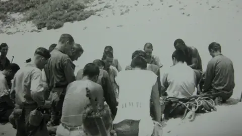 A black and white photograph shows a group of about 20 men sitting and kneeling in prayer on a beach. Some of the men are Japanese, some are American. They all have short hair and have their heads bowed.