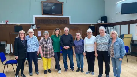 Sally Fairfax Nine people are standing smiling at the camera in a large sports hall with a parquet floor. A wooden carved sign behind them reads "Ferens Hall"
