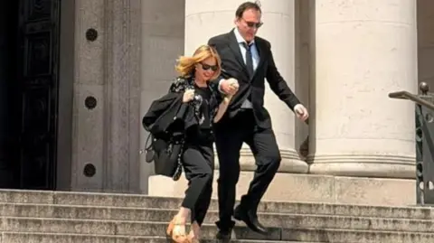 Quentin Tarantino and Kylie Minogue outside National Museum Cardiff. They are walking down the steps outside the museum holding hands. They are both wearing all black outfits and sunglasses. Kylie has a pair of brown tan high heels on.