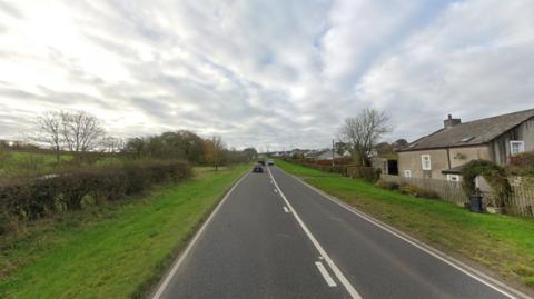 The A595 at Bothel is a rural road. It has grey tarmac, to the left is a hedge with fields in the distance, to the right are some houses.