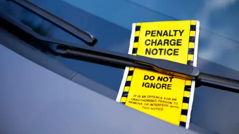 A bright yellow parking fine is tucked under a car's window wiper