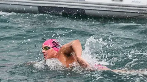William Ellis is swimming in blueish-grey sea besides a grey inflatable support boat. He has a pink swimming cap and is wearing goggles.