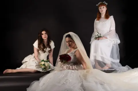 Ashleigh Sunley Three women are modelling white wedding dresses from different eras. The dress on the left looks quite modern and minimalist. The dress in the centre is very intricate and comes with a long veil. The dress on the right is from an earlier style period.   