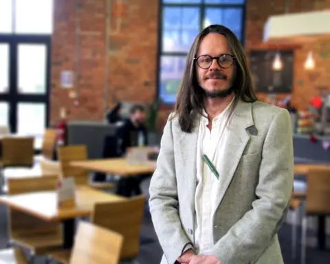 Dr Darren Quelch Darren Quelch, a man with long brown hair in a grey suit and glasses, stands in cafe and looks at the camera