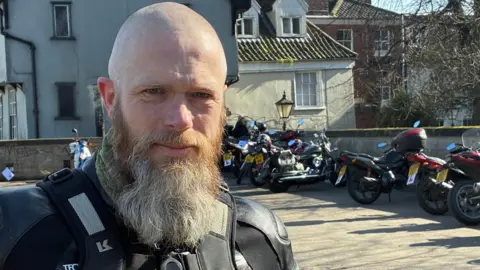A man wearing a black leather jacket stands in front of a row of motorbikes. He has a long grey beard and a shaved head. Behind him, there are buildings.