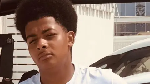 A teenage boy leaning against a car, wearing a white top
